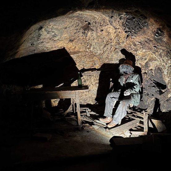 Eine Person sitzt in einem Stuhl in einer Höhle mit einem Tisch, und eine Flasche steht auf dem Tisch.