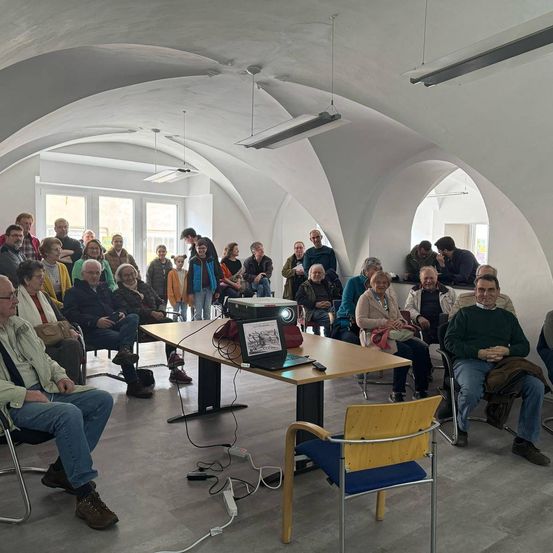 Eine Gruppe von Menschen versammelt sich in einem Saal mit gewölbten Decken und weißen Wänden. Sie sitzen auf Stühlen und scheinen einer Präsentation zuzuhören. Ein Tisch mit einem Beamer und einem Laptop steht vor ihnen.