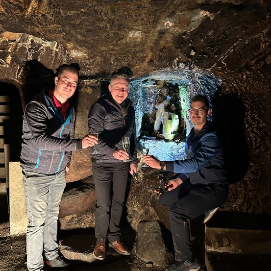Drei Männer stehen in einer Steinhöhle, halten Weingläser in der Hand und lächeln in die Kamera. Hinter ihnen befindet sich eine Wand mit einer Steinstatue.