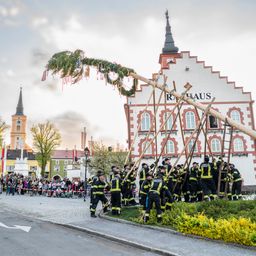 Eine Gruppe von Feuerwehrleuten führt eine traditionelle Aufgabe vor einem Gebäude durch. Eine große Holzkonstruktion wird von mehreren Personen gehalten. Die Szene findet im Freien statt.