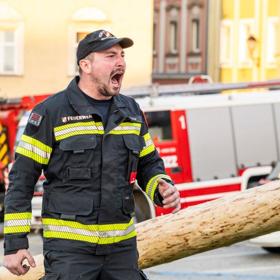 Ein Feuerwehrmann gibt Befehle, während er neben einem Feuerwehrauto steht. Die Szene ist draußen, mit einem gelben Gebäude und einem Baumstamm im Vordergrund.