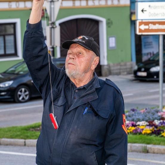 Ein älterer Mann in einem marineblauen Jacken hält ein rotes Werkzeug und steht neben einer Straße mit Straßenschildern und geparkten Autos.