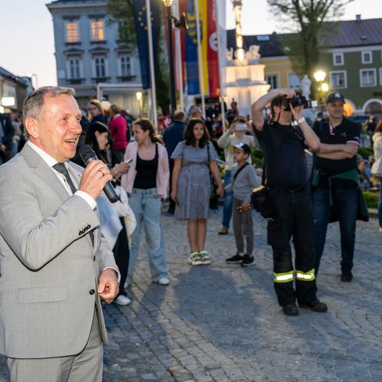 Ein Mann in einem Anzug spricht in ein Mikrofon in einer Außenumgebung. Hinter ihm machen Leute Fotos und stehen herum, darunter zwei Fotografen. Die Szene ist lebhaft mit Zuschauern, und der Hintergrund zeigt Gebäude und Flaggen.