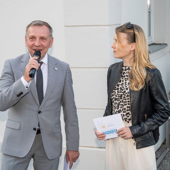 Ein Mann in einem grauen Anzug hält ein Mikrofon und lächelt. Neben ihm hält eine Frau in einer schwarzen Lederjacke ein Stück Papier. Sie stehen vor einem weißen Gebäude.