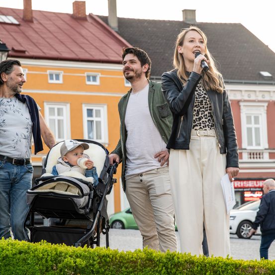 Eine Frau steht und hält ein Mikrofon, möglicherweise hält sie eine Rede, mit einem Mann und einem Baby in einem Kinderwagen neben ihr.
