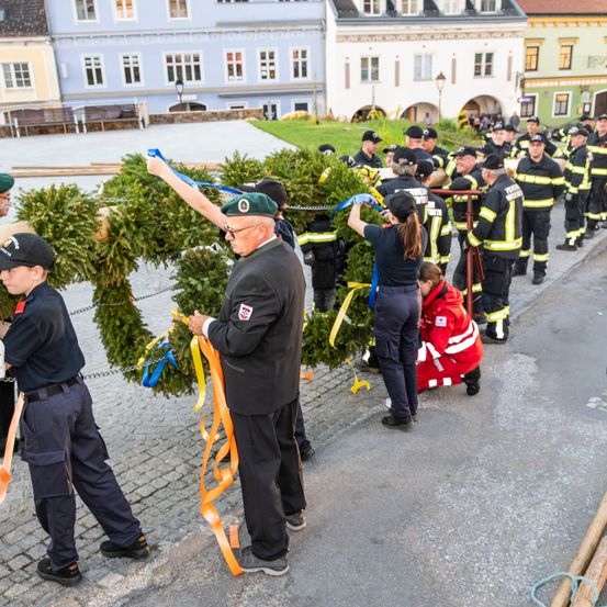 Eine Gruppe von Feuerwehrleuten, einige in Uniform, arbeiten gemeinsam an der Herstellung einer Kranz auf einer Kopfsteinpflasterstraße.