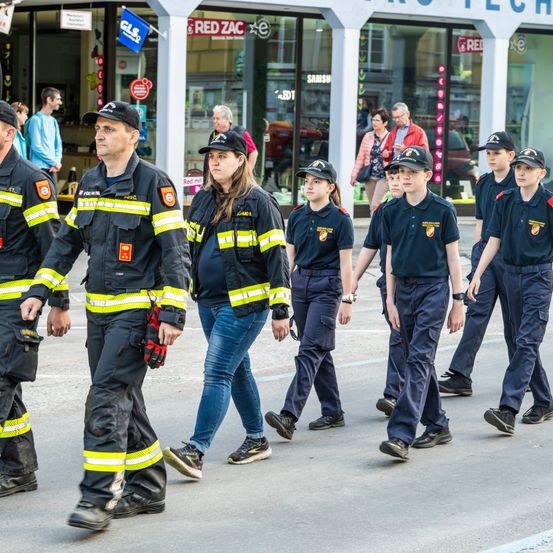 Eine Gruppe von Feuerwehrleuten, darunter Männer und Frauen, läuft die Straße entlang, wahrscheinlich an einer Parade teilnehmend. Sie tragen Uniformen und Mützen. Im Hintergrund gehen Menschen an Geschäften vorbei.