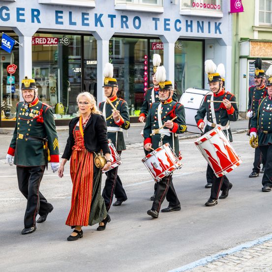 Eine Marschkapelle in traditionellen Uniformen spielt Trommeln auf der Straße, begleitet von einer Frau in einem bunten Kleid.