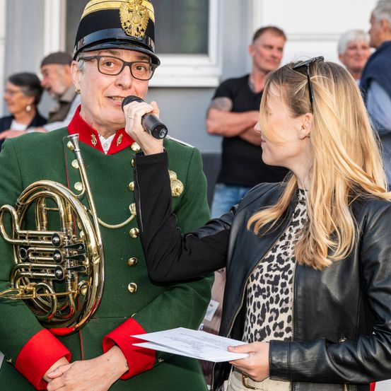 Eine Frau in einer schwarzen Lederjacke hält ein Papier, während ein älterer Mann in einer grünen Militäruniform ein Waldhorn hält und in ein Mikrofon spricht.