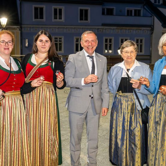 Fünf Personen in traditioneller Kleidung stehen vor einem Gebäude, lächeln und halten Getränke. Ein Mann in Anzug steht in der Mitte.
