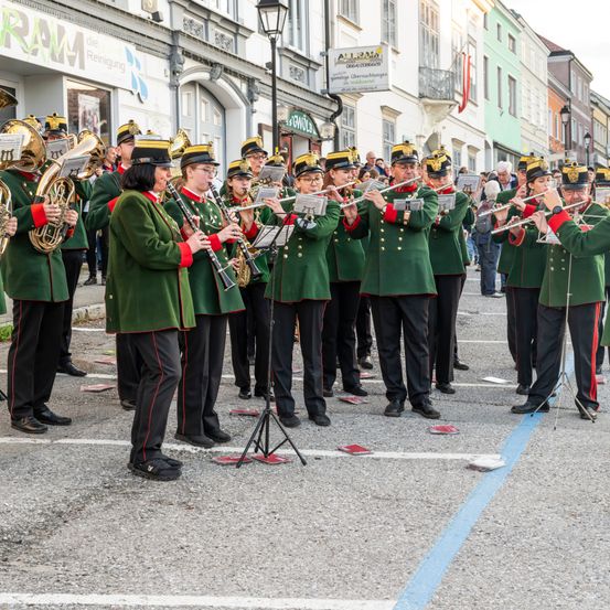 Eine Blaskapelle in grünen Uniformen spielt Instrumente auf der Straße, während einige Leute vom Bürgersteig zuschauen.