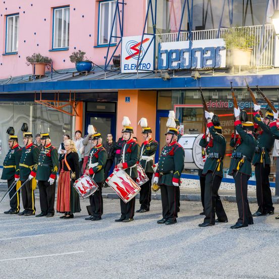 Eine Marschkapelle in grünen Uniformen steht vor einem Geschäft und hält Gewehre und Trommeln. Das Gebäude hat einen blauen Vorbau und Topfpflanzen auf dem Balkon.