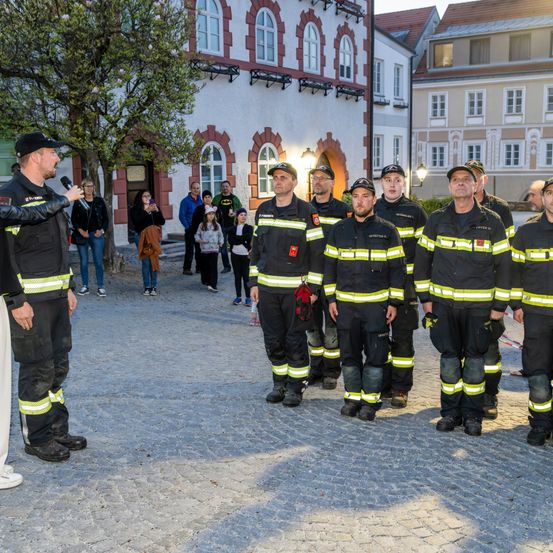 Feuerwehrleute in Uniform stehen vor einem Gebäude, ein Mann spricht in ein Mikrofon. Im Hintergrund gehen und stehen Menschen.