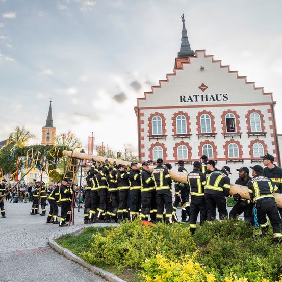 Feuerwehrleute versammeln sich vor dem Rathaus, heben einen großen Baumstamm. Die Menge ist lebhaft, und ein Uhrturm ist im Hintergrund zu sehen.