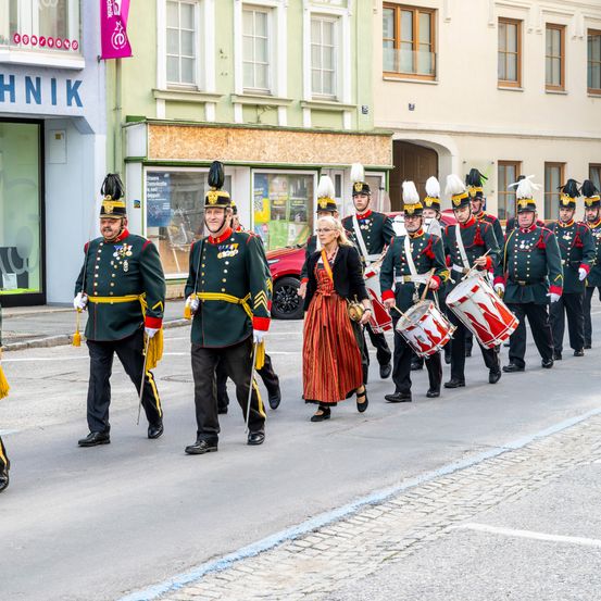 Ein Militärparade auf einer Stadtstraße mit Soldaten in Uniform und einer Frau, die mit einem Trommel vorangeht. Gebäude mit Glasfenstern und einer Fahne im Hintergrund.