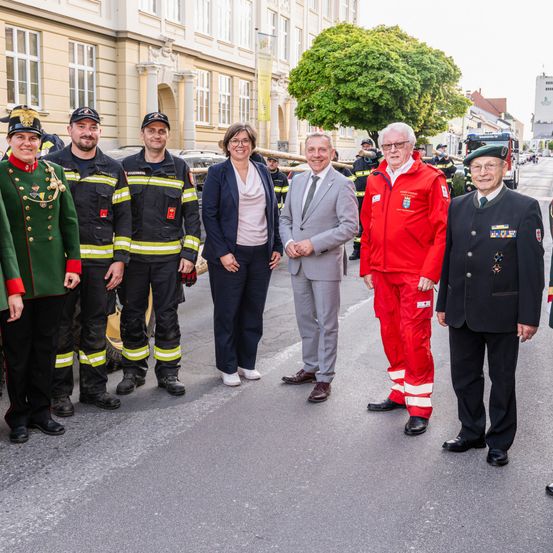 Eine Gruppe von Personen in Uniformen, einschließlich Feuerwehrleuten und Beamten, steht auf einer Straße vor einem Gebäude.