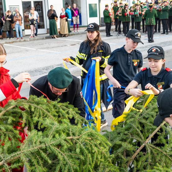Mehrere Personen binden gelbe Bänder um Weihnachtsbäume, mit einer Marschkapelle im Hintergrund.