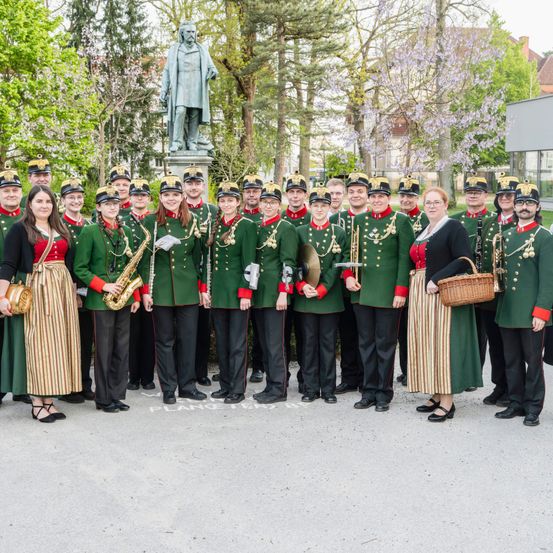 Eine Gruppe von Musikern in grünen Uniformen mit roten Besätzen steht vor einer Statue in einem Park.