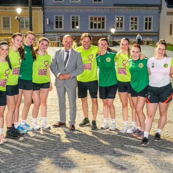 Eine Gruppe junger Frauen in grünen Shirts und Shorts posiert mit einem Mann im Anzug vor einem Gebäude.