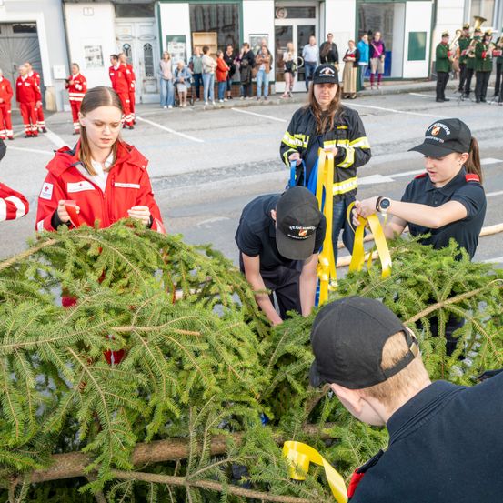 Eine Gruppe von Menschen schmückt einen Tannenbaum, einige in roten Uniformen und andere in Schwarz. Sie binden gelbe Bänder an den Baum.