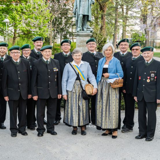 Eine Gruppe älterer Personen in traditioneller Kleidung, einschließlich Hüten und Medaillen, posiert für ein Foto im Park.