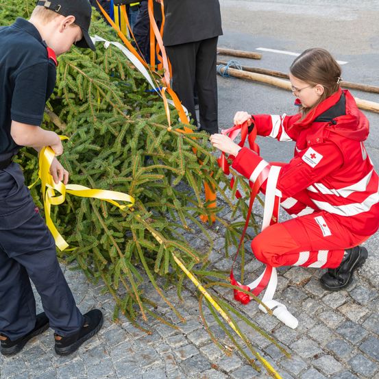 Zwei Personen dekorieren einen Tannenbaum mit gelben Bändern, während eine dritte Person hinter ihnen steht.