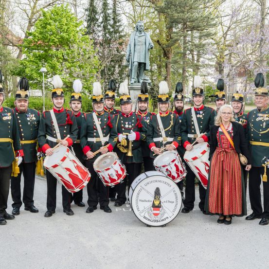 Eine Gruppe von Männern in grünen Uniformen posiert mit Trommeln vor einer Statue. Eine Frau in traditioneller Kleidung steht in der Nähe.