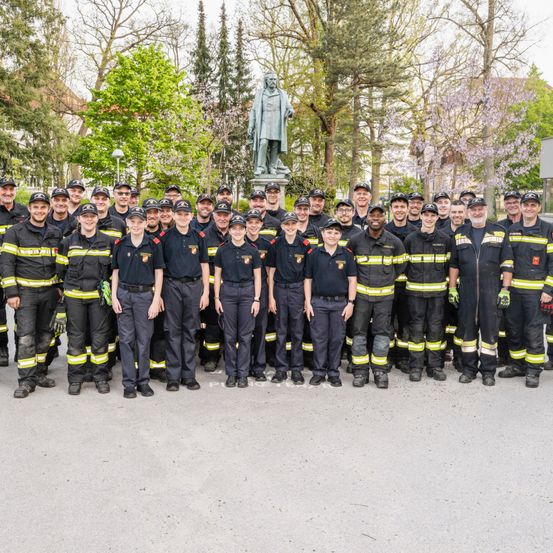 Eine Gruppe Feuerwehrleute in Uniform steht in einem Park, mit einer Statue im Hintergrund.