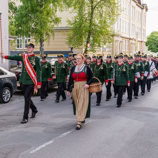 Ein Marschkapelle in grünen Uniformen geht eine Stadtstraße hinunter, angeführt von einem Mann in einem zeremoniellen Schal. Eine Frau mit einem Korb folgt. Gebäude und geparkte Autos säumen die Straße.