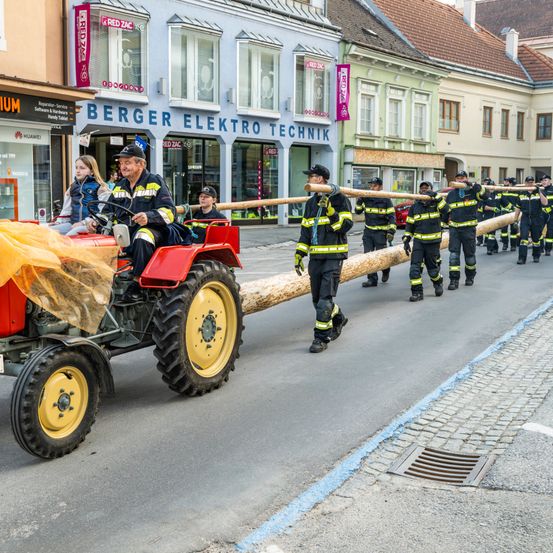 Ein Traktor mit Feuerwehrleuten fährt durch eine Straße mit Geschäften, darunter ein RED ZAC-Laden. Feuerwehrleute laufen auch mit einem großen Baumstamm.
