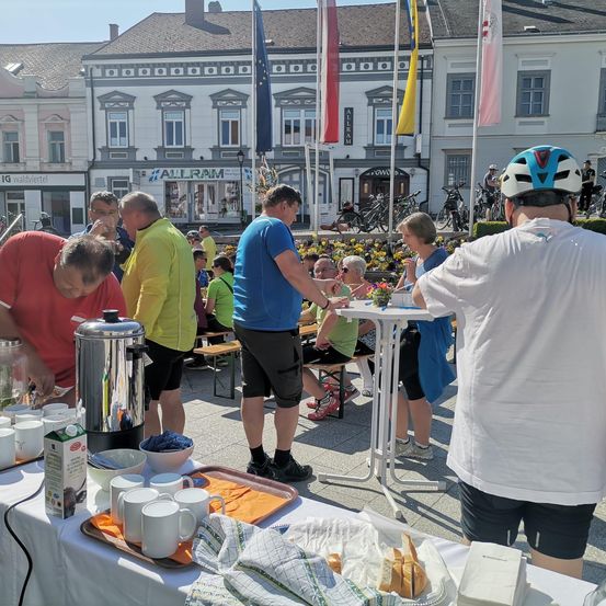Men essen und trinken an einem Tisch draußen auf der Straße. Ein Mann trägt einen weißen Helm.