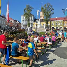 Eine Gruppe von Radfahrern sitzt an Tischen in einem Außenbereich. Einige von ihnen tragen Helme. Es stehen und gehen Leute an der Seite. In der Ferne befindet sich ein Denkmal mit einer Statue, Bäumen und Gebäuden.