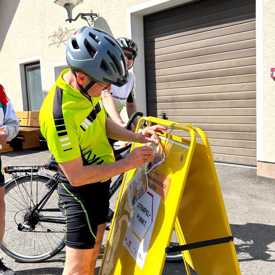 Ein Mann in einem gelben Fahrradtrikot platziert einen Aufkleber auf einem gelben Schild, das 'Stempel-Punkt' sagt. Er trägt einen Helm. Eine andere Person steht neben ihm mit einem Fahrrad. Eine Lampe ist an der Wand montiert, und eine Holzbank ist auf der linken Seite platziert.