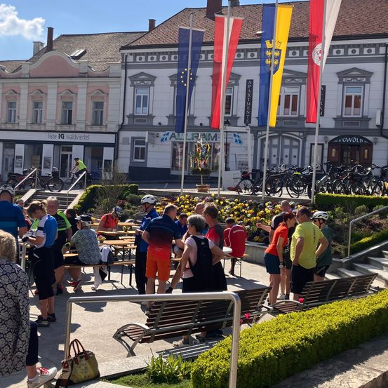 Menschen versammeln sich vor einem Gebäude mit vielen Fahnen, in der Nähe einer Bank und eines Fahrradparkplatzes. Eine Person fährt auf dem Bürgersteig mit dem Fahrrad.