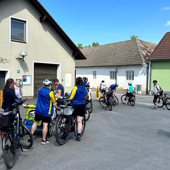 Eine Gruppe von Radfahrern mit Helmen bereitet sich auf ihre Fahrt vor. Sie stehen auf einem Parkplatz versammelt, mit mehreren Häusern im Hintergrund.