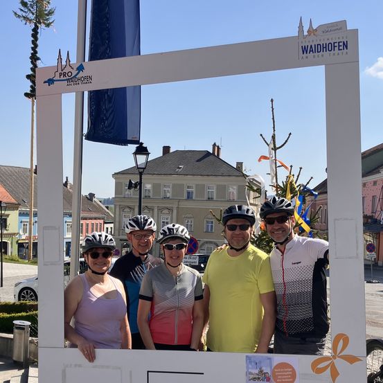 Fünf Menschen in Helmen und Sonnenbrillen posieren für ein Foto vor einem weißen Rahmen mit einem Logo darauf. Dahinter steht eine blaue Fahne, ein Laternenpfahl und Gebäude.