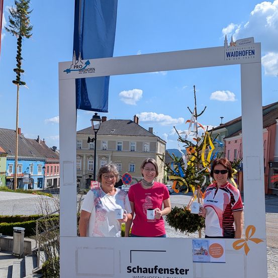 Drei Frauen stehen in einer Fotobox mit Tassen in den Händen. Die Box ist mit einem Weihnachtsbaum dekoriert und hat den Text 'Schaufenster'. Dahinter stehen eine Fahne, eine Straßenlaterne und Gebäude.