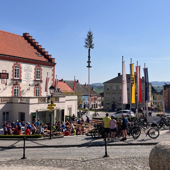 Eine Gruppe von Menschen versammelt sich auf einem Kopfsteinpflasterplatz, einige sitzen an Tischen, während andere stehen und herumlaufen. Fahrräder sind in der Nähe geparkt und verschiedene Flaggen sind an Stangen gehisst. Der Platz ist von Gebäuden umgeben und in der Ferne sind Hügel zu sehen.