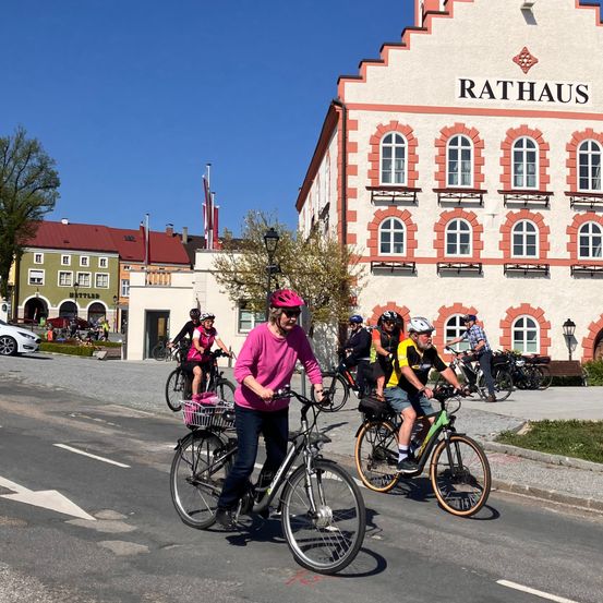 Eine Gruppe von Menschen fährt auf einer Straße mit Fahrrädern vorbei an einem Gebäude mit der Aufschrift 'Rathaus', das ein rotes Dach und viele Fenster hat, unter einem klaren Himmel.