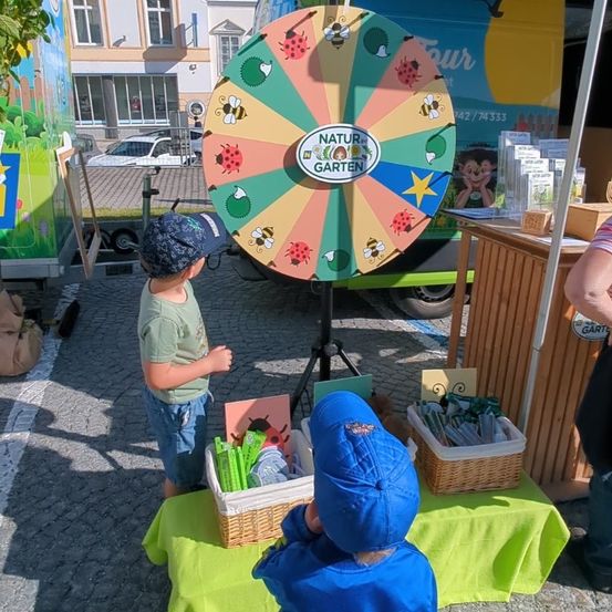 Zwei Kinder stehen bei einem großen Drehrad an einem Gartenstand. Ein Kind trägt ein grünes Shirt und einen blauen Hut, das andere ein blaues Jacket und einen Hut. Sie betrachten das Rad, auf dem verschiedene Insekten abgebildet sind. Vor ihnen stehen Körbe mit Gegenständen auf dem Tisch.