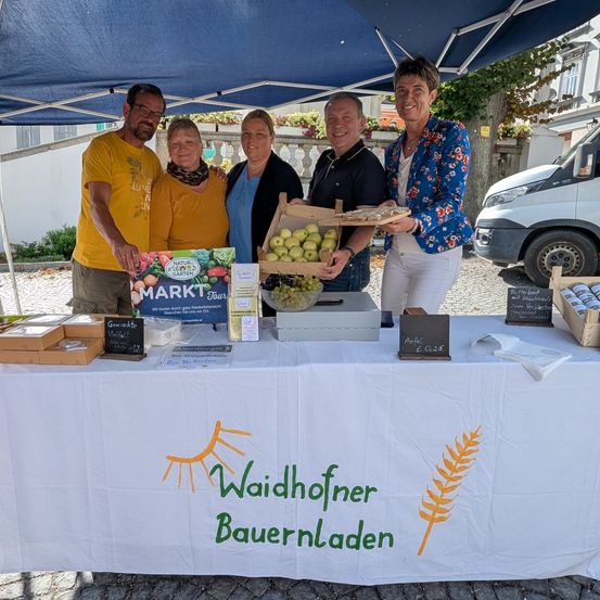 Fünf Menschen stehen lächelnd unter einem Zelt. Ein Tisch hat Kisten, Äpfel und ein Schild mit der Aufschrift 'Waidhofer Bauernladen.' Dahinter steht ein Van.