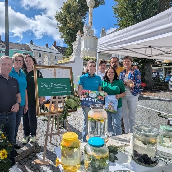 Mehrere Personen stehen hinter einem Tisch mit Gläsern gefüllt mit Flüssigkeiten, einige halten ein Buch. Sie befinden sich in einem Außenbereich, mit einem weißen Baldachin über ihnen. Eine Statue steht hinter ihnen, und Gebäude sind im Hintergrund.