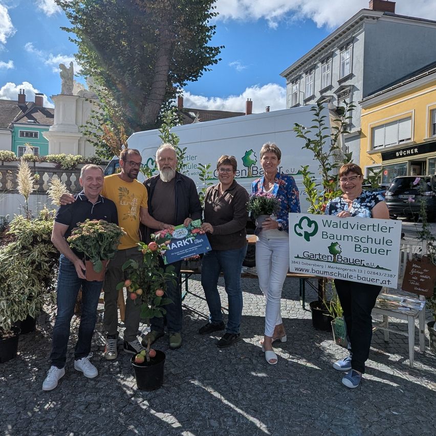 Fünf Personen stehen zusammen auf einem Stadtplatz mit Pflanzen und einem Banner. Sie tragen Freizeitkleidung und haben Blumen im Vordergrund. Dahinter ist ein weißer Lieferwagen und Gebäude zu sehen.