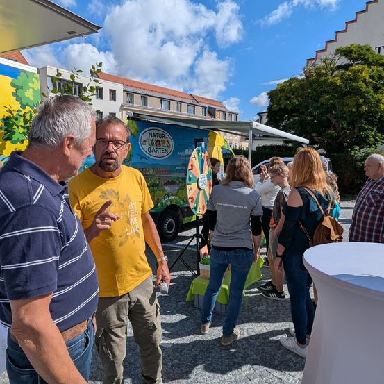 Eine Outdoor-Veranstaltung, eine Gruppe von Menschen diskutiert etwas. Ein Mann in einem gelben Shirt zeigt auf etwas, während andere zuhören. Ein Truck mit einem farbenfrohen Design steht hinter ihnen.