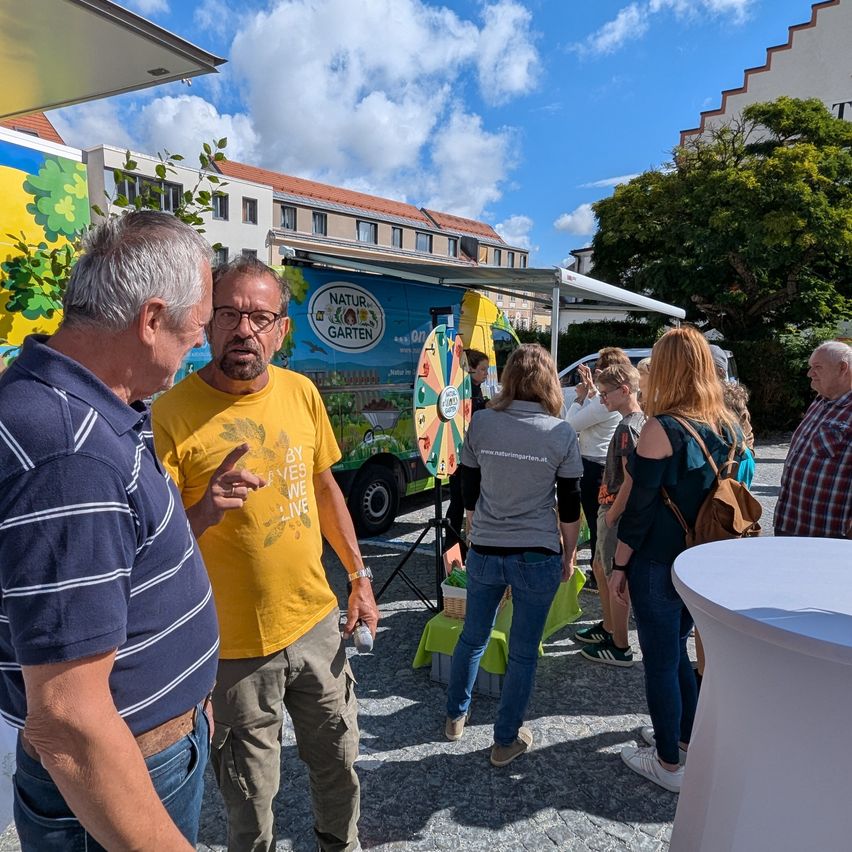 Eine Outdoor-Veranstaltung, eine Gruppe von Menschen diskutiert etwas. Ein Mann in einem gelben Shirt zeigt auf etwas, während andere zuhören. Ein Truck mit einem farbenfrohen Design steht hinter ihnen.