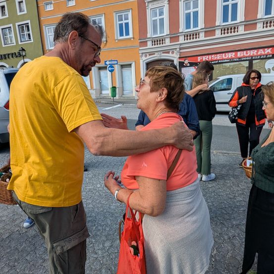 Ein erwachsener Mann und eine Frau umarmen sich im Freien. Der Mann trägt ein gelbes T-Shirt und die Frau ein rosa Oberteil. Sie sind von mehreren Menschen umgeben.