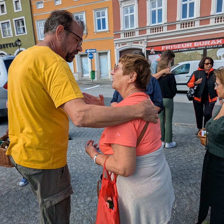 Ein erwachsener Mann und eine Frau umarmen sich im Freien. Der Mann trägt ein gelbes T-Shirt und die Frau ein rosa Oberteil. Sie sind von mehreren Menschen umgeben.
