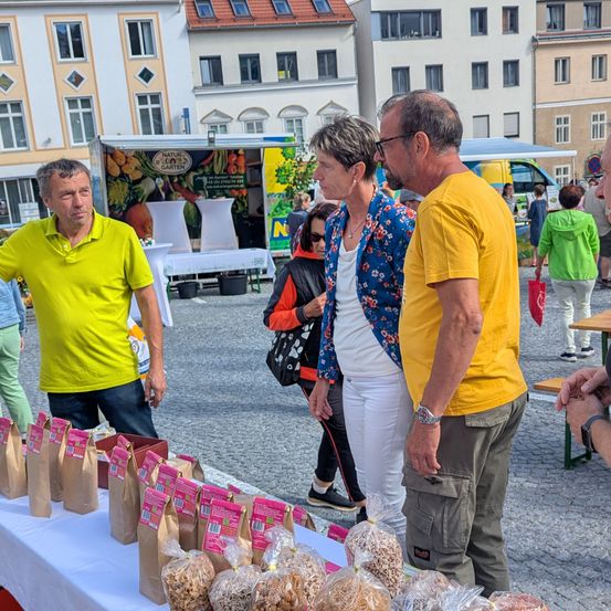 Eine Gruppe von Menschen auf einem Markt mit einem Tisch voller Lebensmittel in Tüten. Ein Mann in einem gelben Shirt spricht mit einer Frau in Blumenkleidung. Ein weiterer Mann in einem gelben Shirt beobachtet.