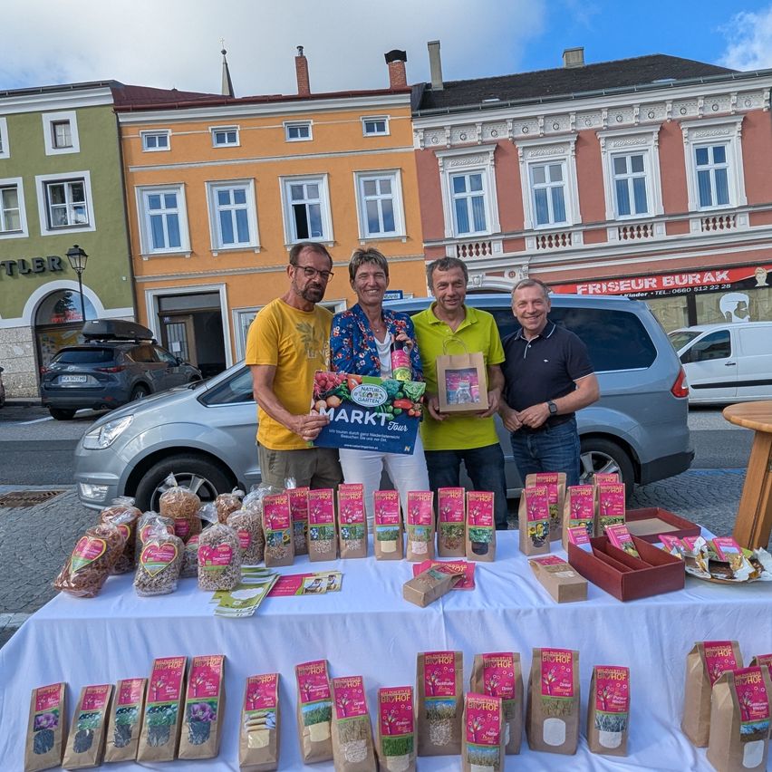 Drei Männer stehen vor einem Tisch mit Säcken voller Samen und einem Schild, das Markt Tour sagt. Sie lächeln und scheinen für ein Foto zu posieren. Der Tisch ist mit einem weißen Tuch bedeckt. Hinter ihnen stehen Gebäude, ein Auto und ein Lieferwagen.