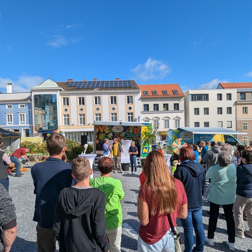 Eine Gruppe von Menschen versammelt sich um einen Imbisswagen, wahrscheinlich bei einer Veranstaltung, unter einem blauen Himmel mit einigen Wolken. Ein Gebäude mit Solarpanelen steht hinter ihnen.